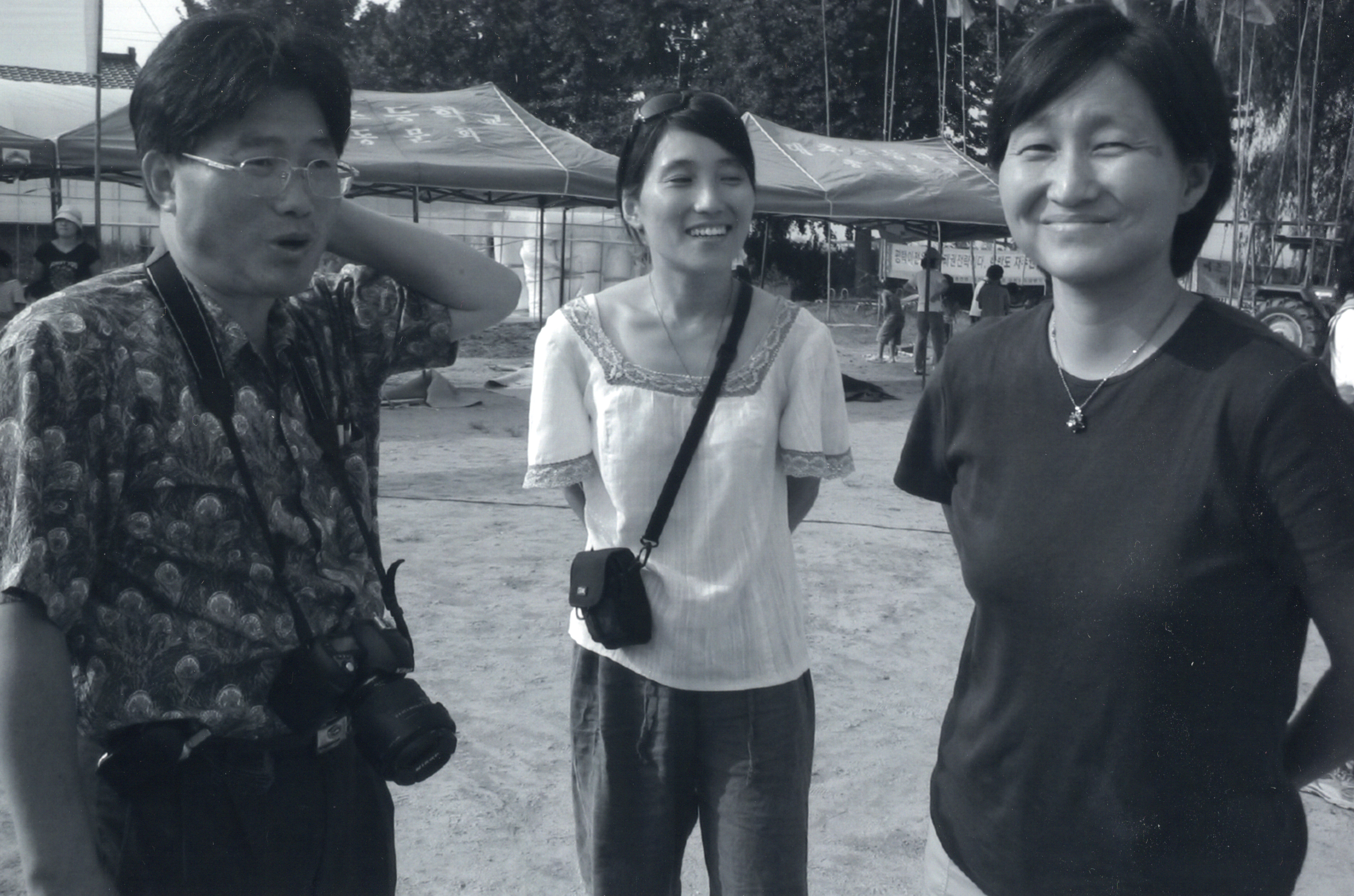 Three Korean people smiling and standing together. there are some tents in the background.