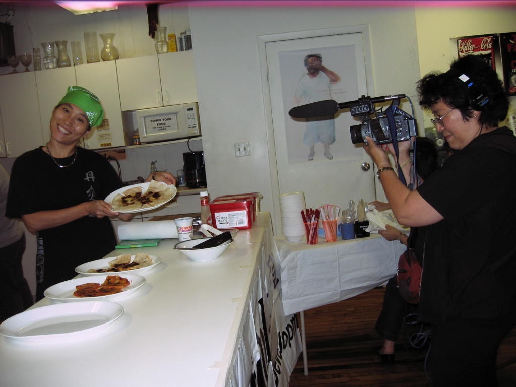 Hyun wearing a green bandana and holding up a plate of food. Someone else in all black is smiling and filming them.