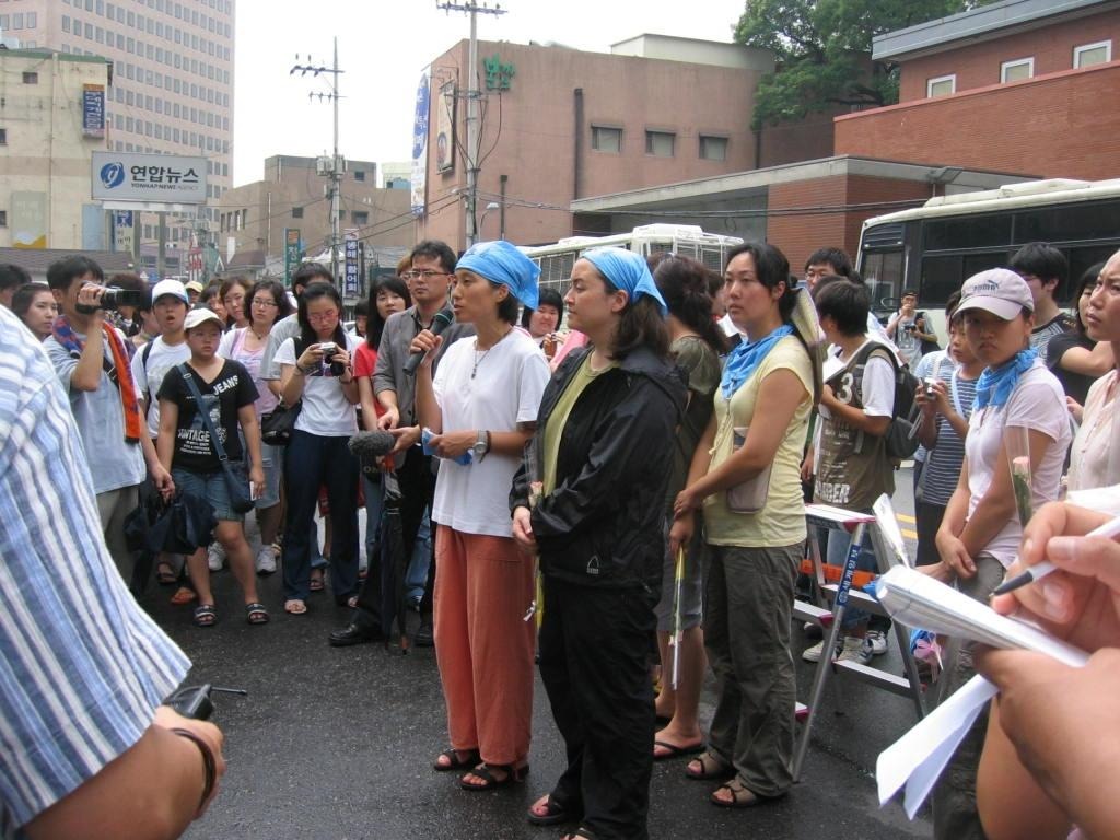 Hyun, younger, with a small group of people all wearing blue bandanas. Hyun is giving a speech in south Korea in front of a larger crowd.