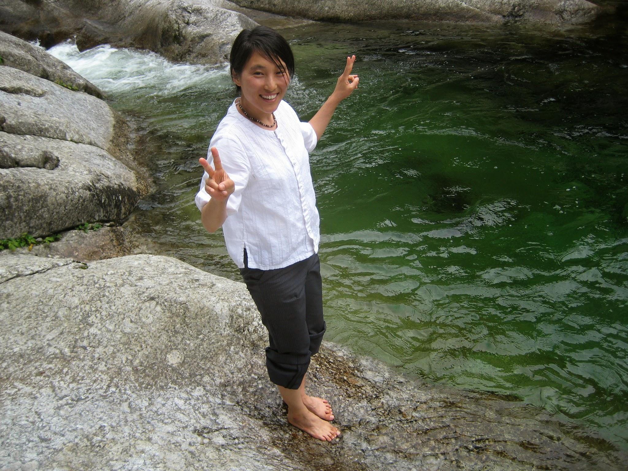 hyun in a white shirt and brown pants, smiling, and holding up peace signs. She is standing in the water in a creek.