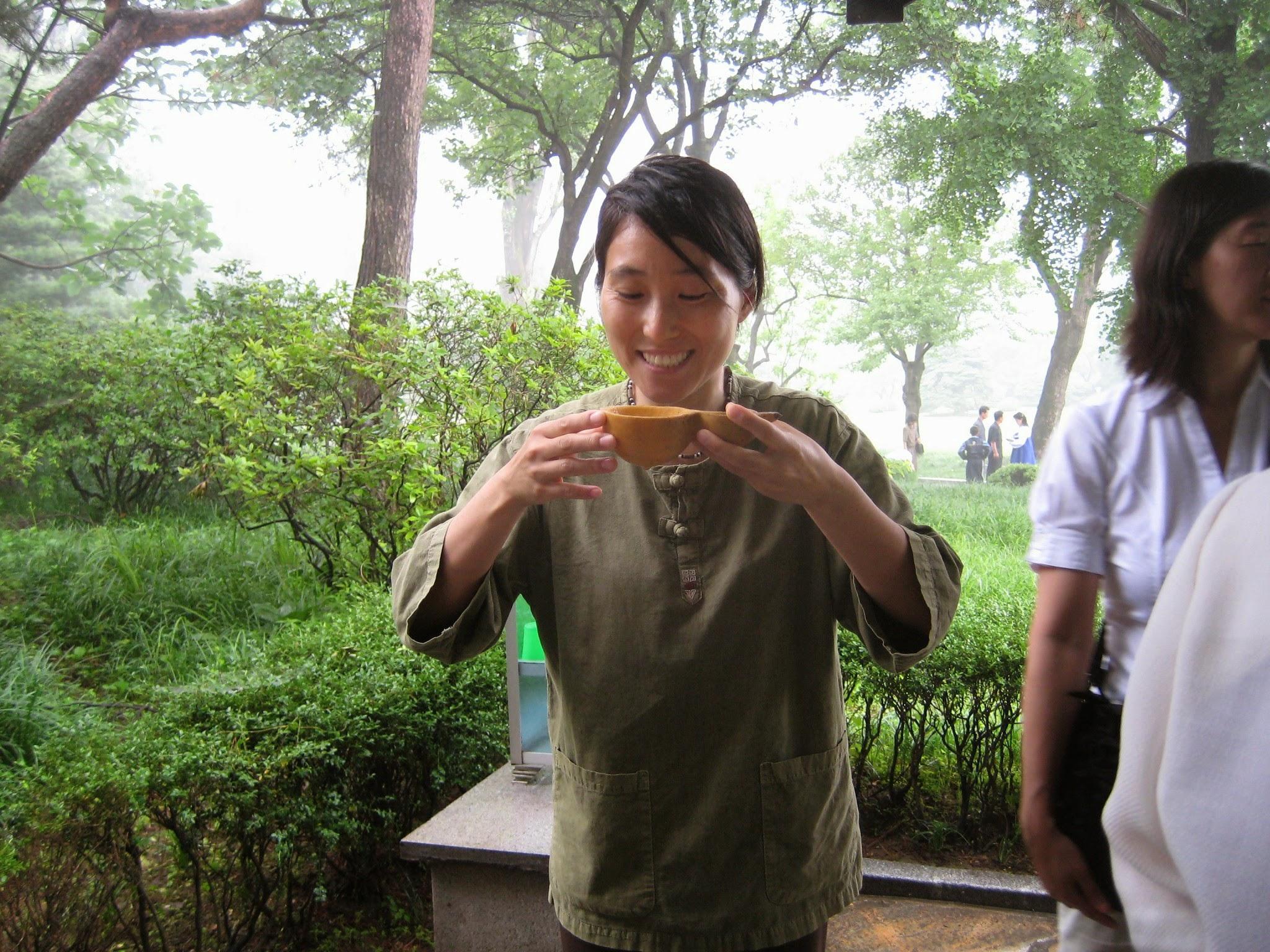 Hyun in the woods holding up a bowl made of squash full of water. She is smiling and about to drink the water.