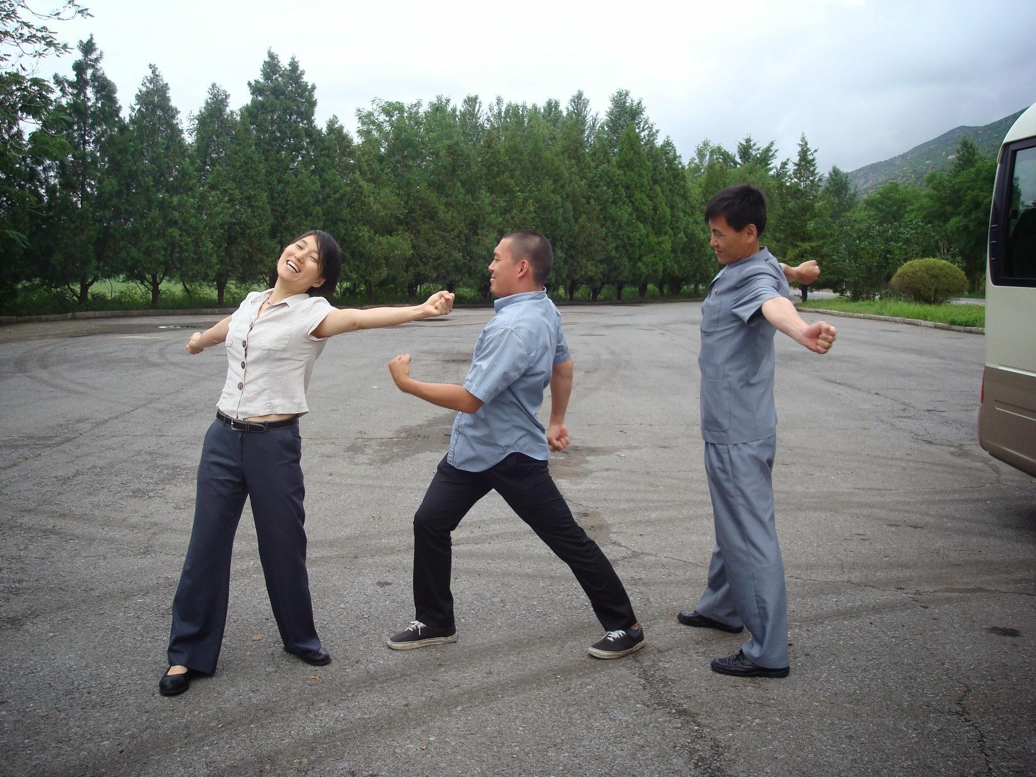 Three Korean people are stretching and dancing, and smiling in a parking lot. Greenery int he background.