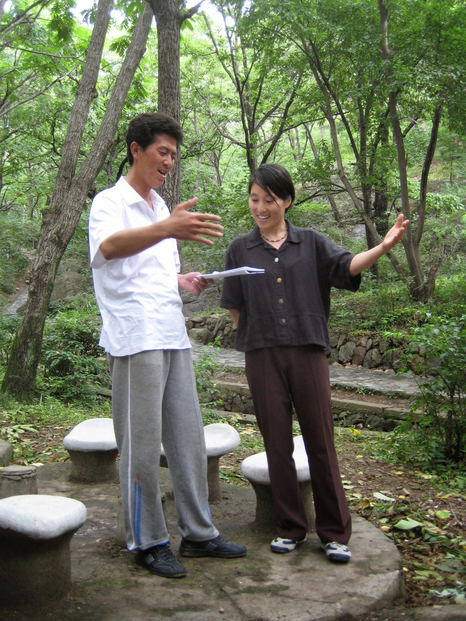 Hyun and a man singing in the woods in the mountains with their arms out in emphasis. They are smiling.