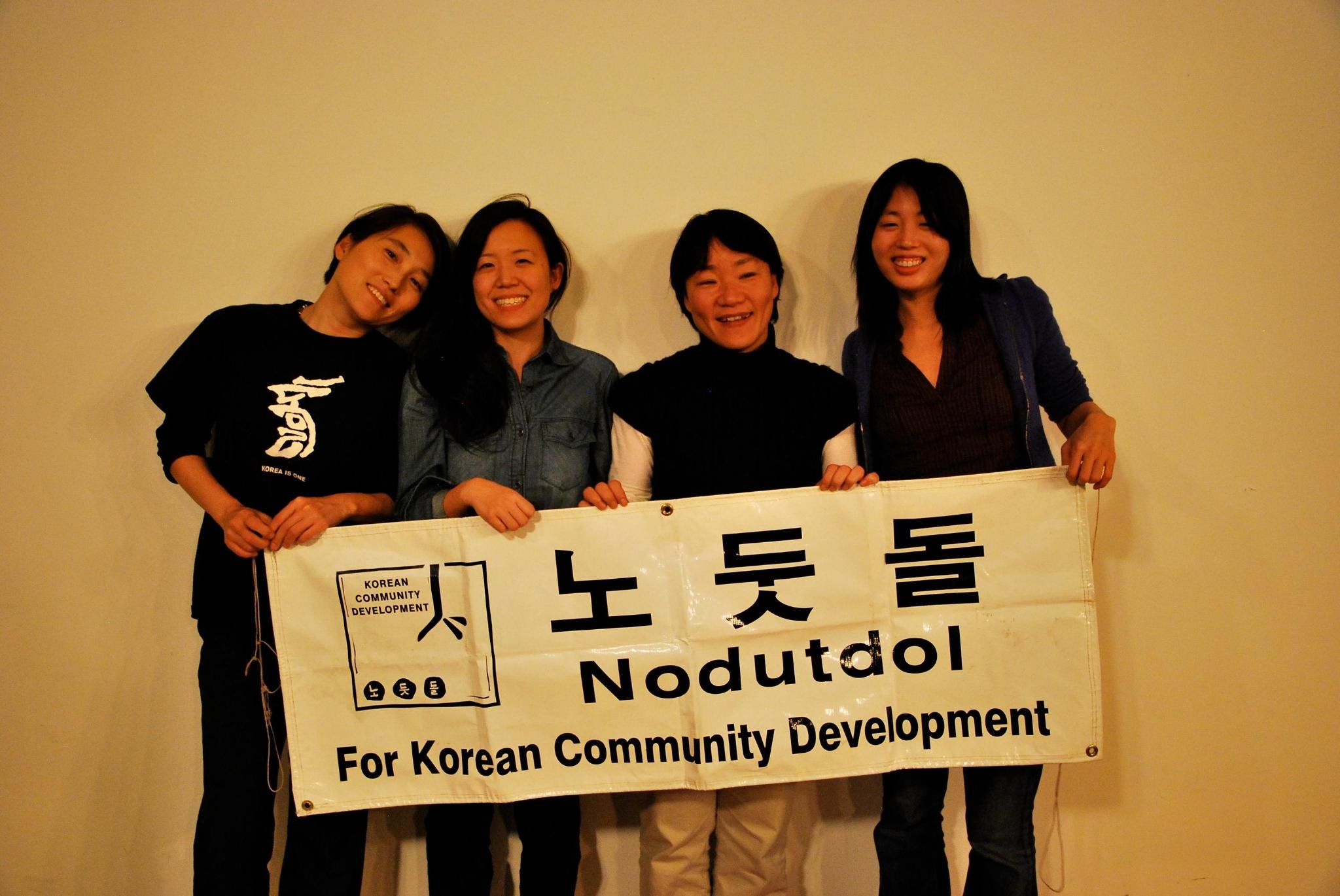 Four Korean women hold up a Nodutdol banner. They are smiling and looking into the camera.
