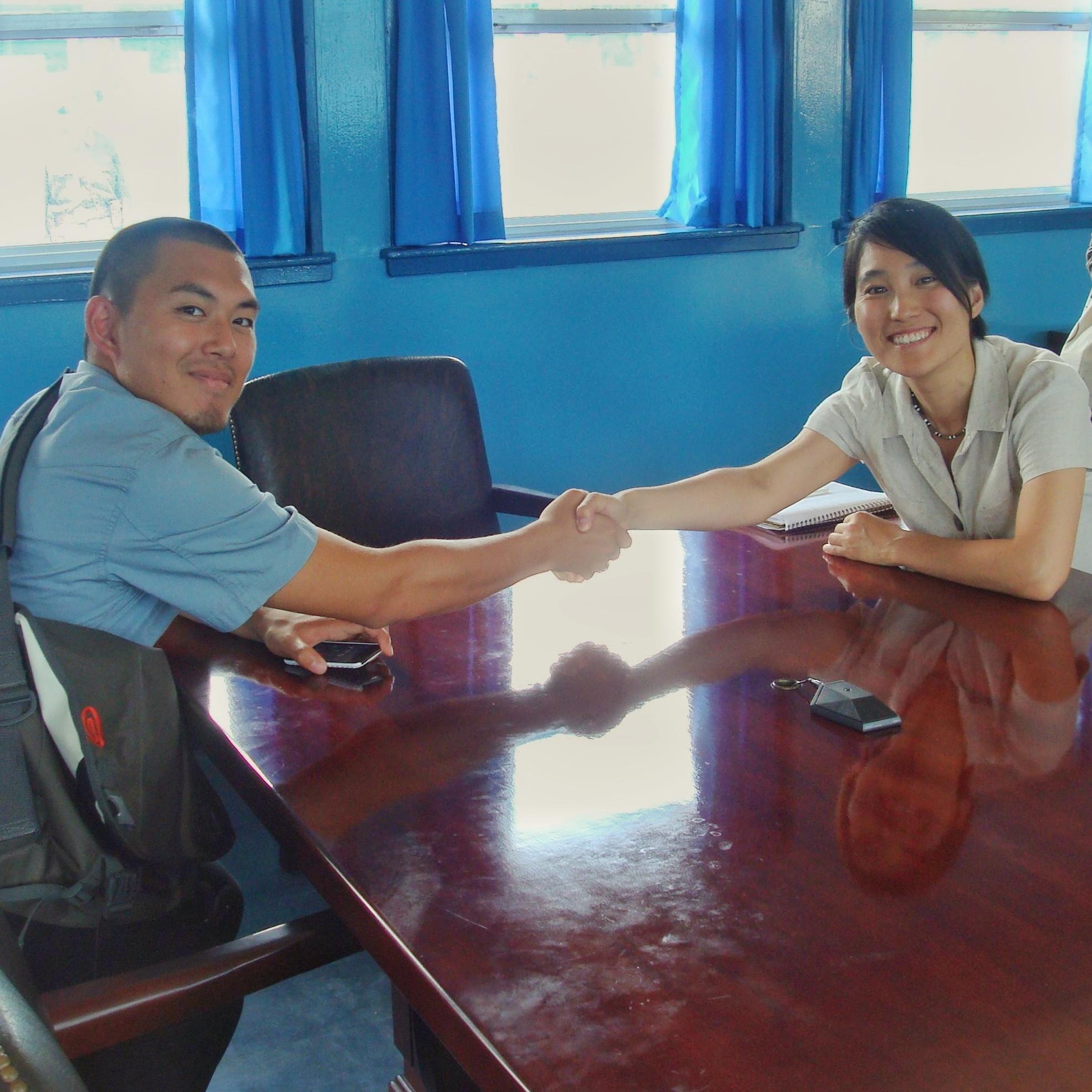 Two Korean people sit at a desk and hold hands across the desk at a blue building at the DMZ.