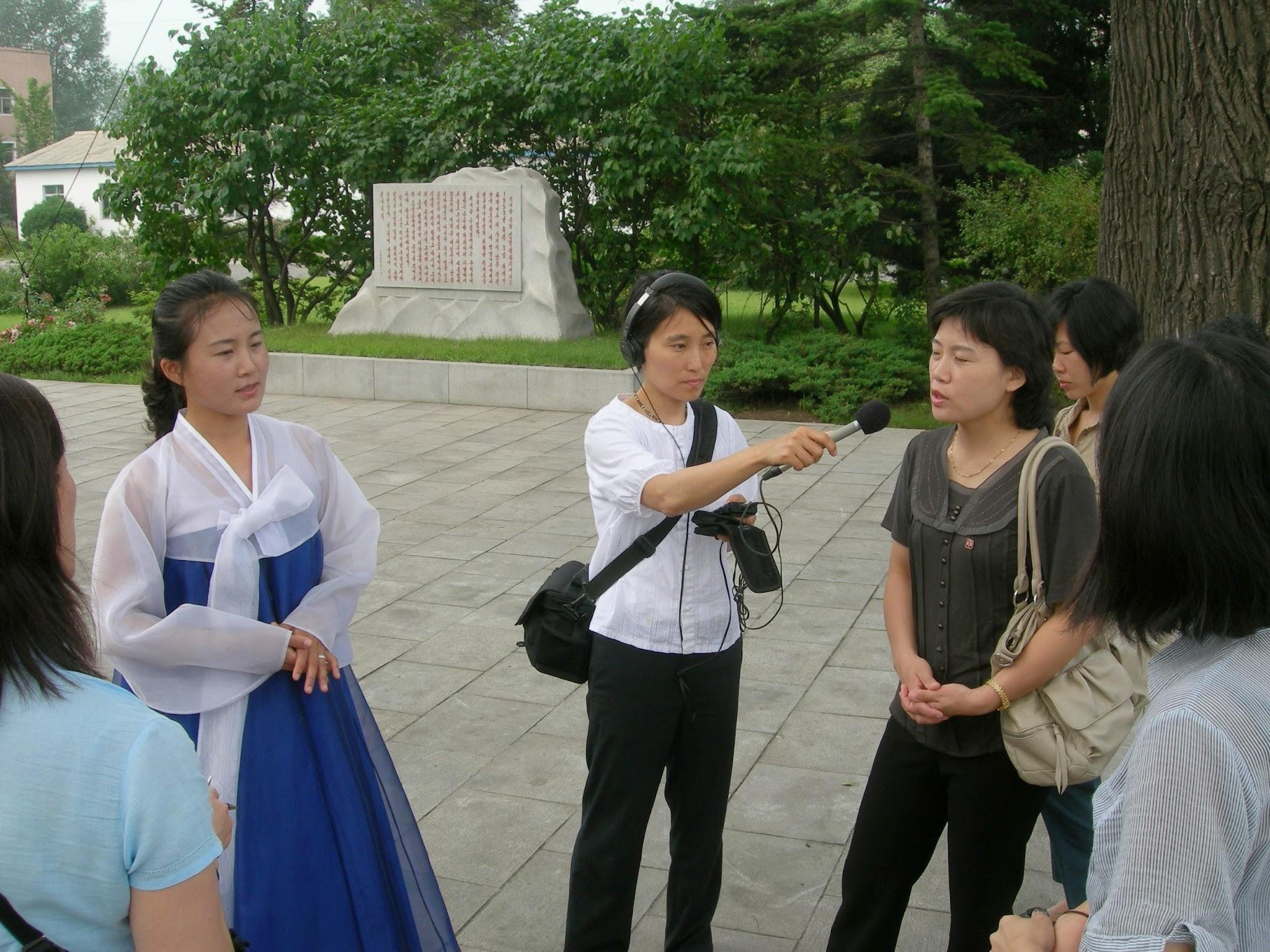 Hyun is wearing white and is wearing some audio gear and recording another woman in north Korea. There are other women surrounding them and they are standing in front of a small monument.